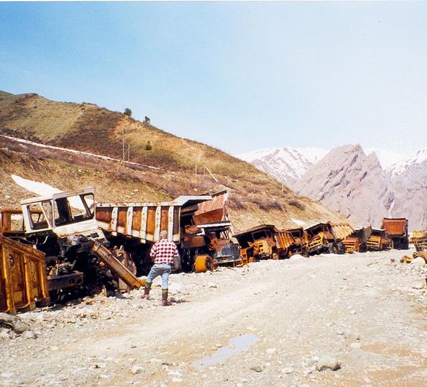 burned dump trucks below a hill. A man in plaid surveys the damage